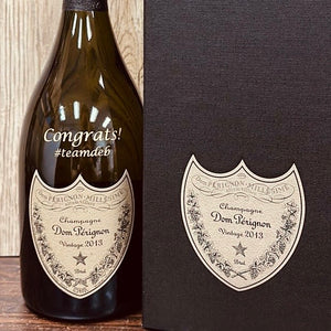 A wine bottle with a custom engraved label that reads 'Congrats! #teamwed.' The label is attached to the front of the bottle, which is placed against a wooden background.